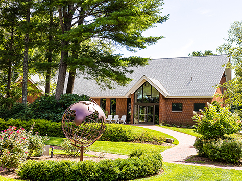 A brick building with a peaked glass entrance behind a garden and a copper globe sculpture.